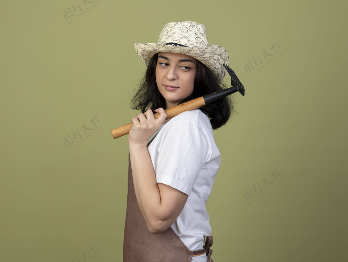 绿色年轻的黑发女园丁穿着制服 戴着园艺帽 站在一旁耙橄榄色帽子