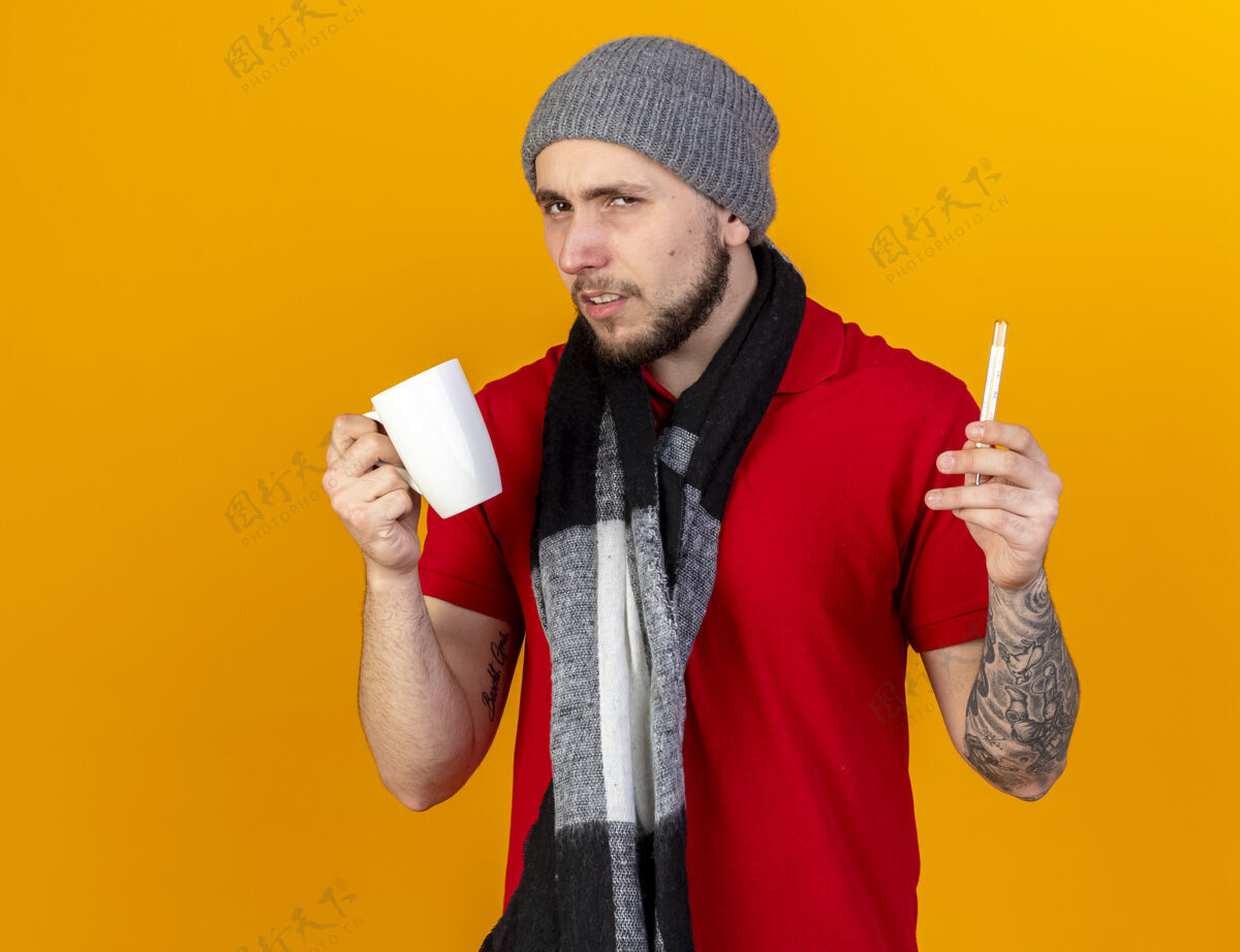 疾病一个戴着冬帽和围巾的年轻白种人 手里拿着橘子色的杯子和温度计冬天围巾穿