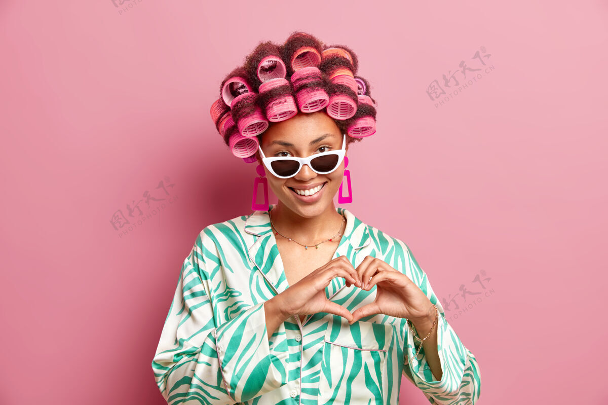 形状相当满意的美国黑人妇女的水平镜头应用卷发器做发型穿丝绸睡衣太阳镜和耳环形状的心标志隔离在粉红色的墙上脸人类手势