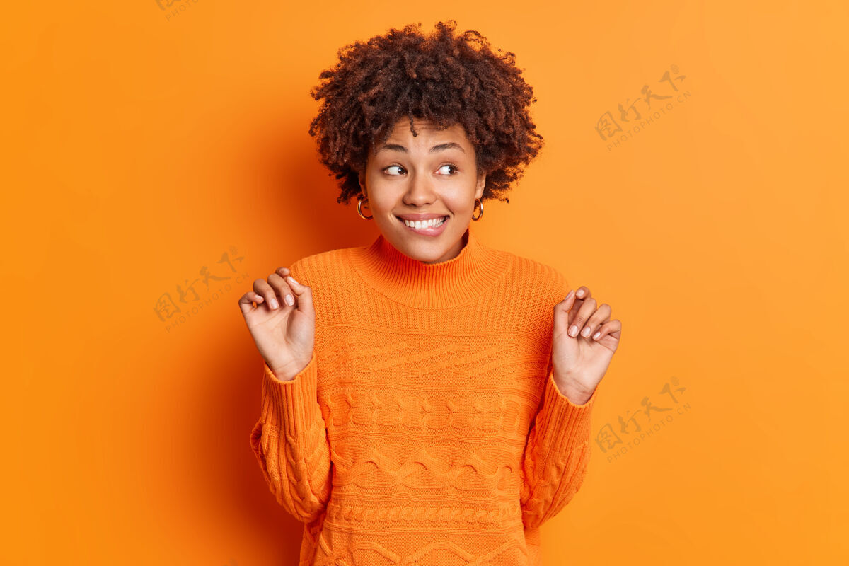 乐观半长镜头正面的非裔美国妇女咬嘴唇保持举手好奇地看着远处微笑好奇地注意到一些有趣的东西穿休闲套头衫在橙色的墙上满足好奇女人