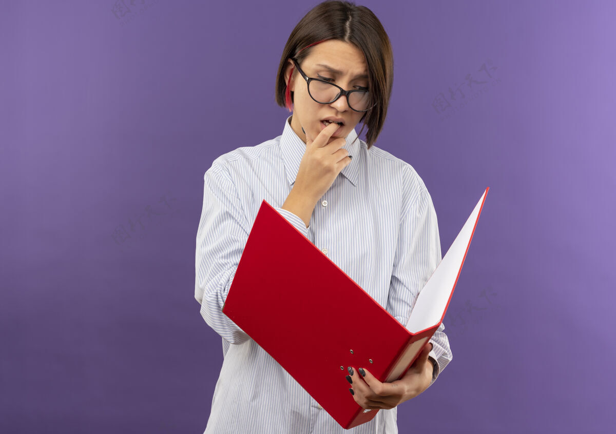 呼叫体贴的年轻呼叫中心女孩戴着眼镜 拿着文件夹看着 咬着她的手指孤立在紫色的墙上姿势手指衣服