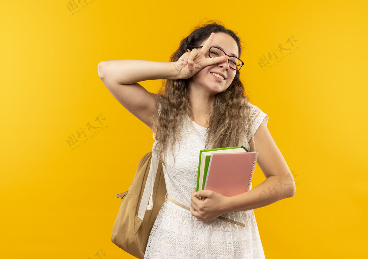 黄微笑着的年轻漂亮的女学生戴着眼镜 背着书包 拿着笔记本 做着隔离在黄色墙上的和平标志抱人年轻
