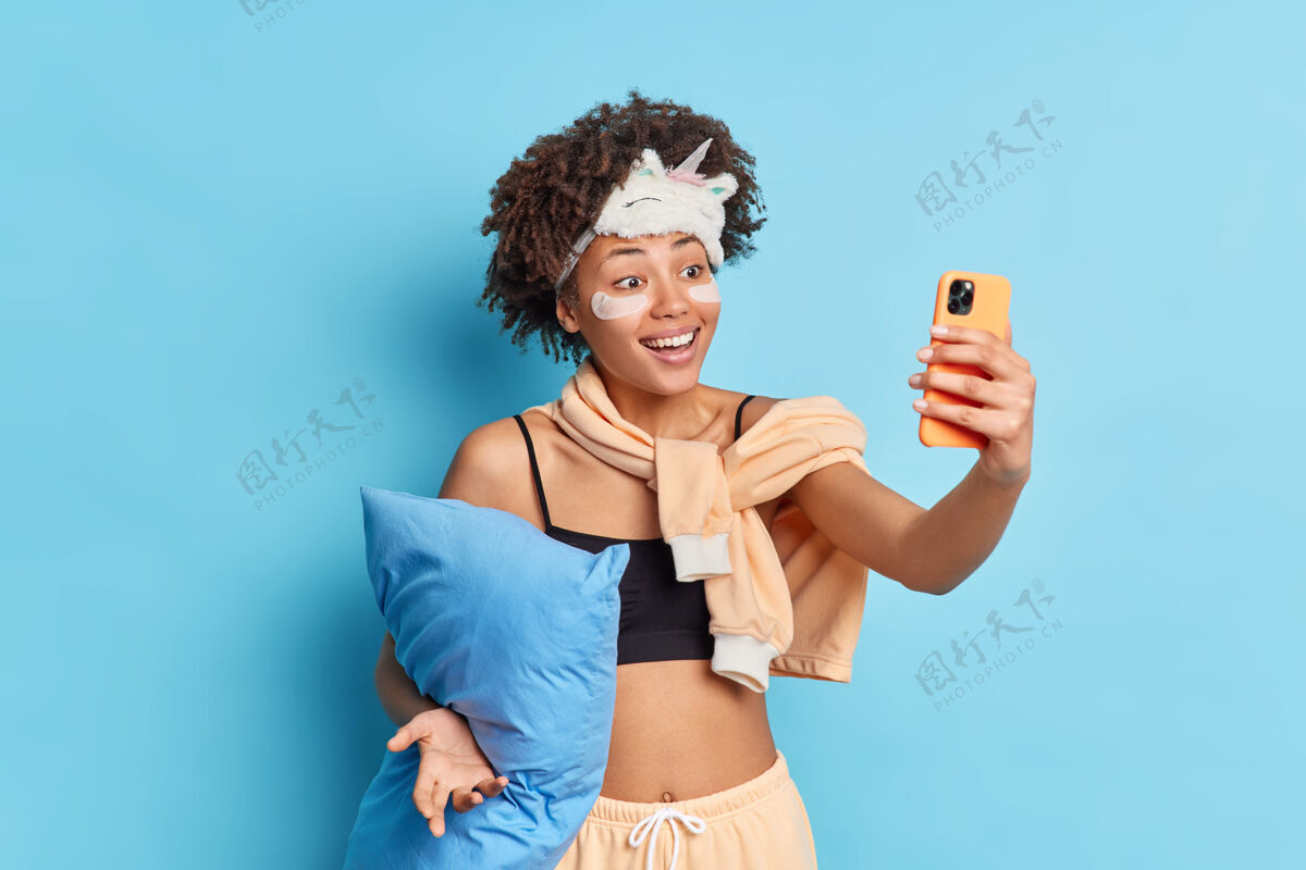 醒着积极的卷发年轻的非裔美国妇女通过智能手机自拍微笑愉快地将胶原蛋白贴在眼睛下面穿着睡衣抱枕隔离在蓝色的工作室墙上室内短信睡衣