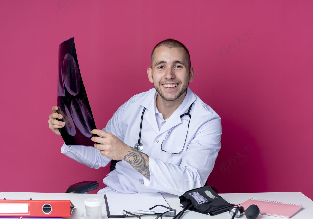 长袍面带微笑的年轻男医生 穿着医用长袍 听诊器 坐在办公桌旁 手里拿着工作工具 把x光照片隔离在粉红色的墙上人穿着人