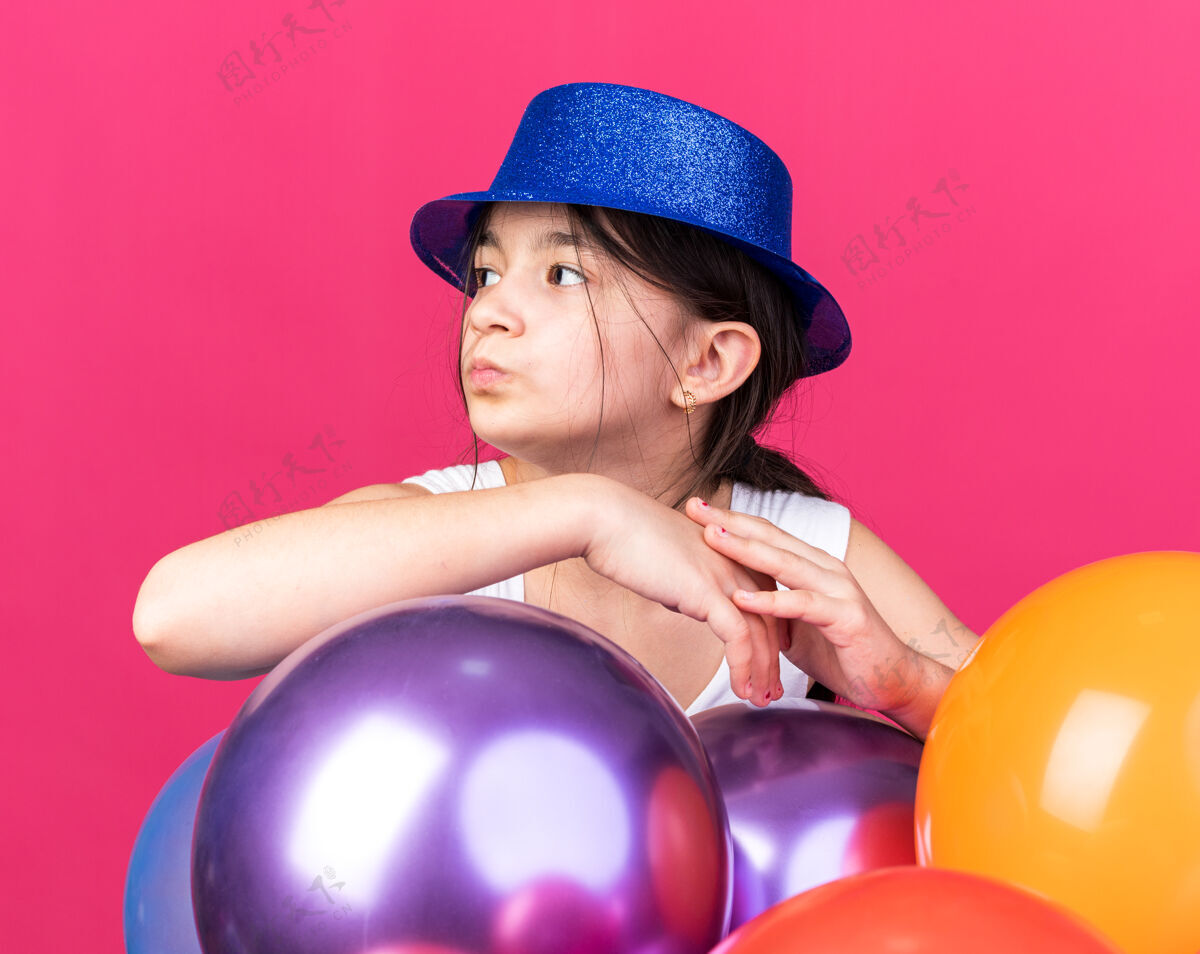 帽子自信的年轻白人女孩戴着蓝色的派对帽 站在氦气球旁边 看着粉色墙壁上孤立的一面 还有复制空间气球自信白种人
