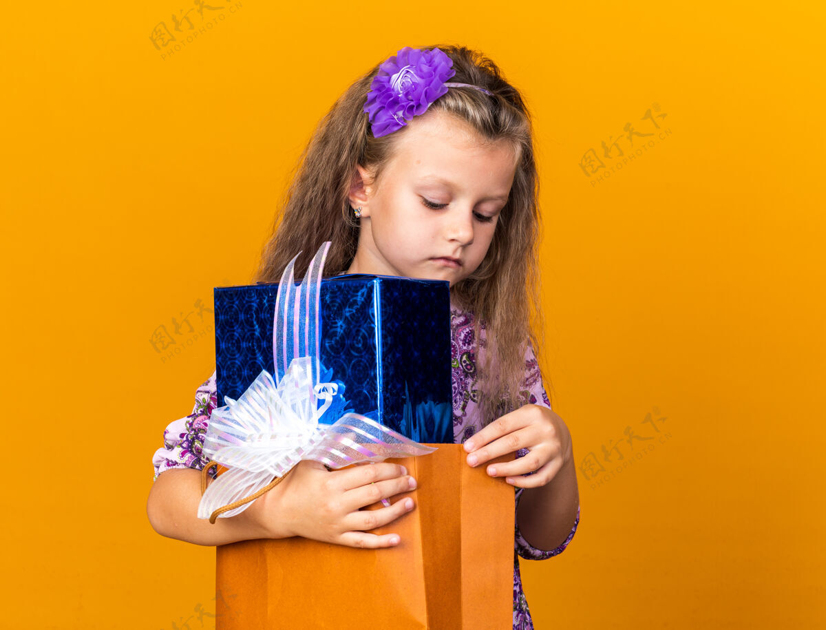 小自信的金发小女孩拿着礼物盒 看着橙色的墙壁上孤立的复制空间金发抱着孤立