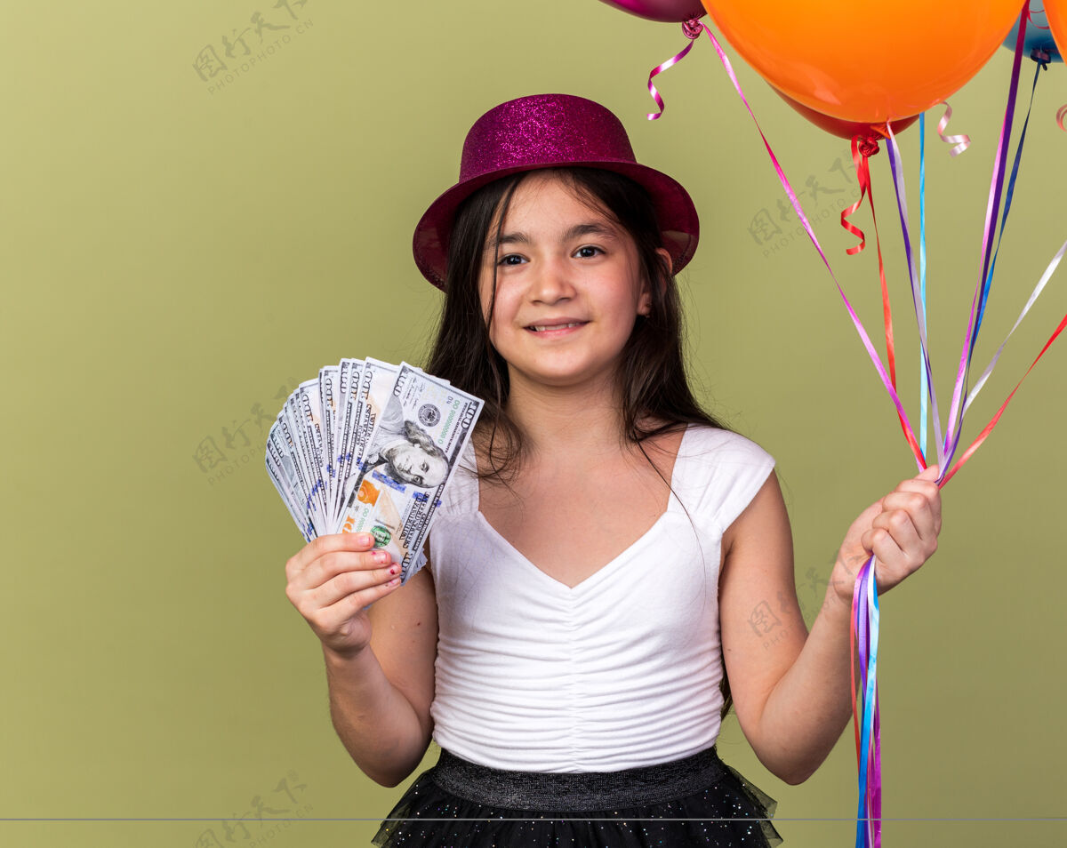 生日微笑着的年轻白人女孩 戴着紫色派对帽 手里拿着钱 氦气球孤立在橄榄绿的墙上 还有复制空间氦年轻橄榄