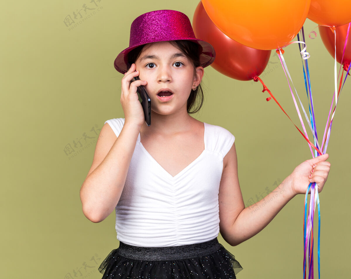 女孩震惊的年轻白人女孩 戴着紫色派对帽 手里拿着氦气球 在橄榄绿的墙上 隔着一块空白的地方讲电话气球氦白种人