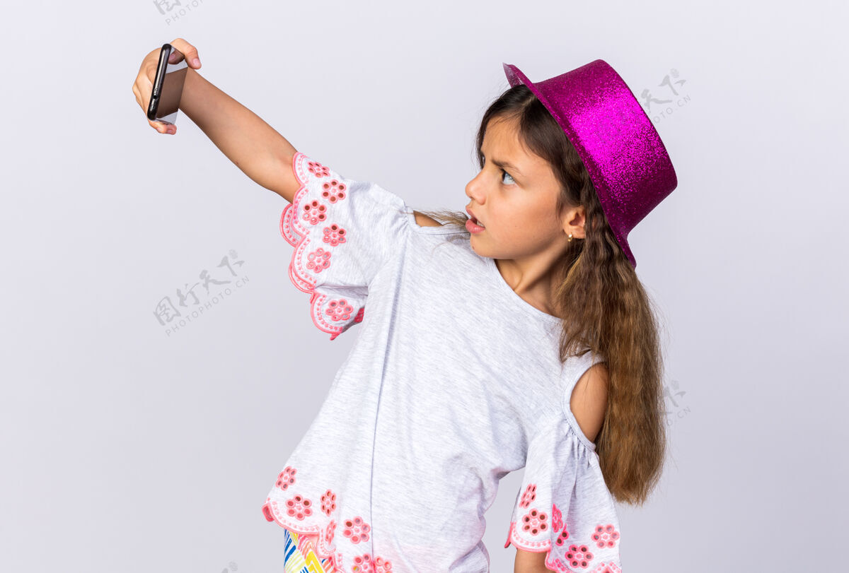 聚会惊讶的小白种女孩戴着紫色的派对帽看着手机在白色的墙上自拍 还有复制空间生日采取帽子
