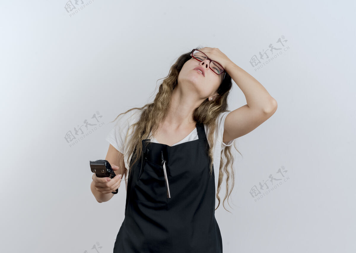 无聊年轻的专业美发师 围着围裙 手里拿着修剪器 手放在额头上 站在白色的墙上又累又无聊衣服人女人