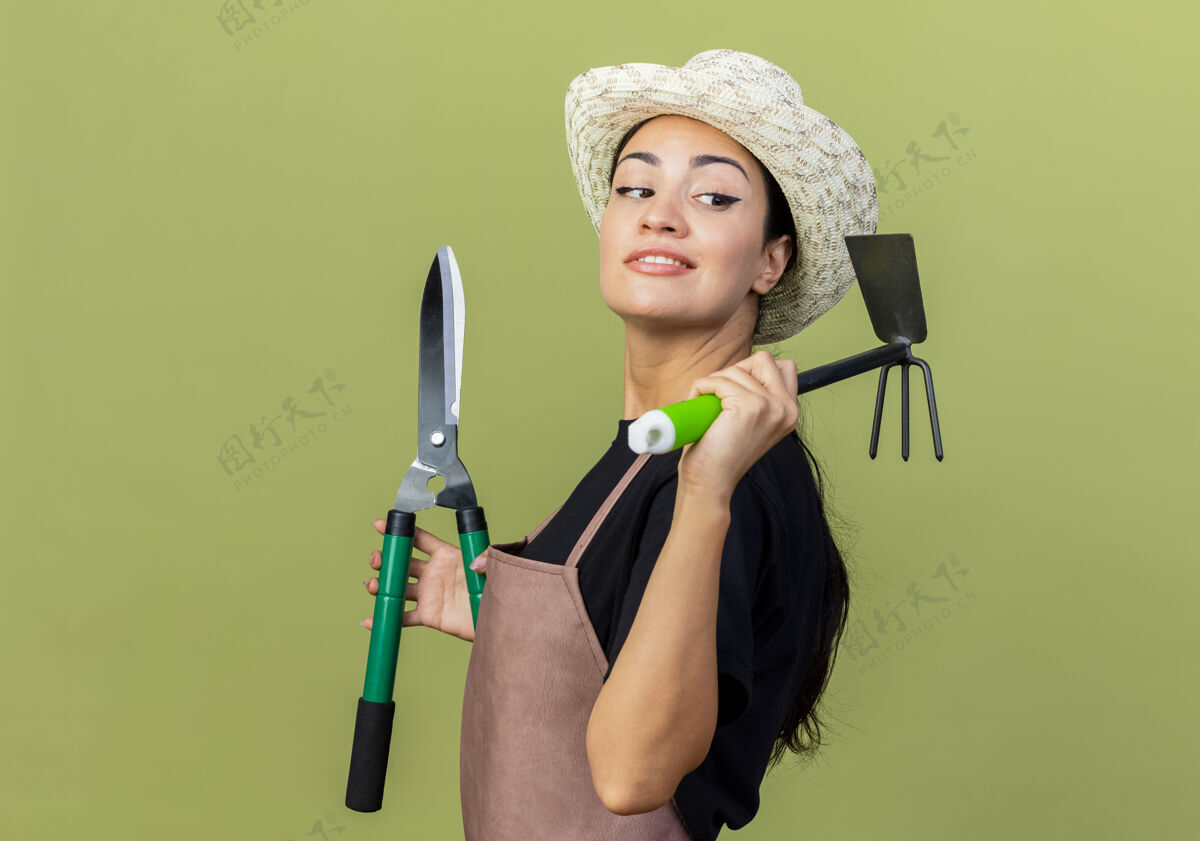 市民年轻漂亮的女园丁 围着围裙 戴着帽子 拿着树篱剪和床垫 站在浅绿色的墙边微笑着向一旁望去微笑床垫女人