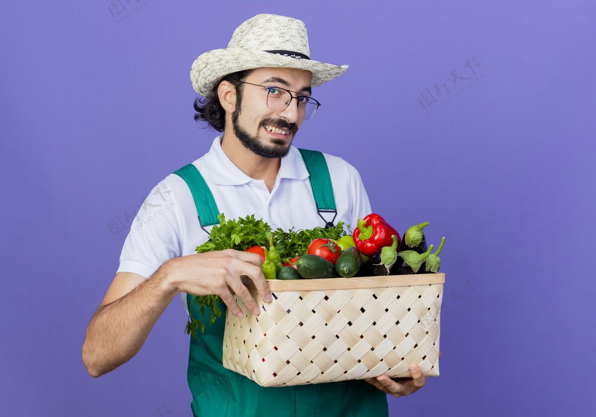 脸年轻的留着胡须的园丁 穿着连体衣 戴着帽子 手里拿着装满蔬菜的箱子 站在蓝色的墙上 面带微笑地看着前方市民微笑蔬菜