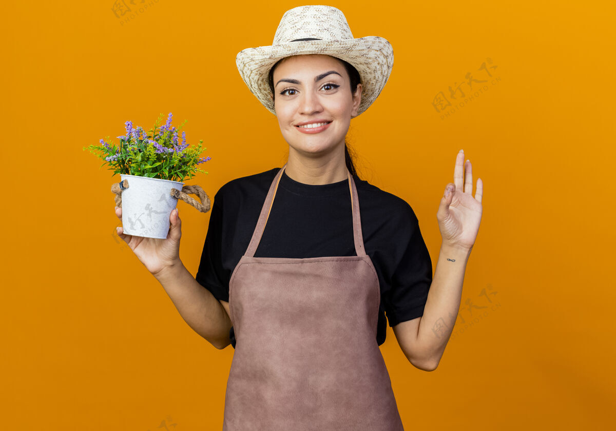 漂亮年轻漂亮的女园丁围着围裙 戴着帽子 手里拿着盆栽植物 微笑着看着前面 橙色的墙上挂着“ok”的牌子感觉市民年轻