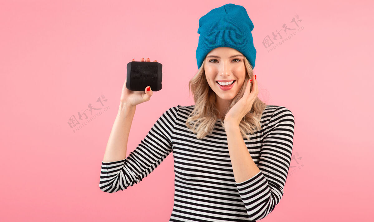 模特年轻女子手持无线扬声器听音乐穿着条纹衬衫和蓝色帽子微笑着摆出粉红色的姿势乐趣音频享受