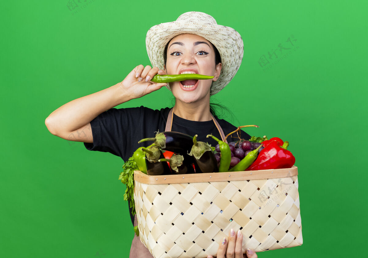 人年轻漂亮的女园丁围着围裙 戴着帽子 手里拿着装满蔬菜的篮子 咬着青椒站在绿色的墙上咬辣椒花园