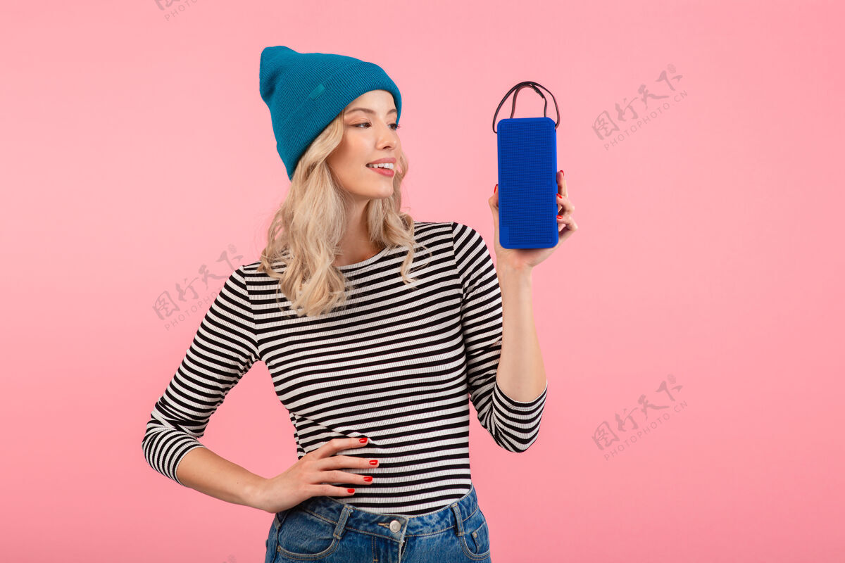无线年轻女子手持无线扬声器听音乐穿着条纹衬衫和蓝色帽子微笑着摆出粉红色的姿势快乐粉色乐趣