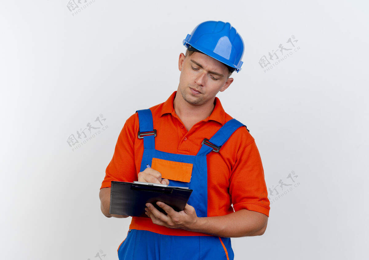 白色想着年轻的男建筑工人穿着制服 戴着安全帽在剪贴板上写些什么头盔制服思考