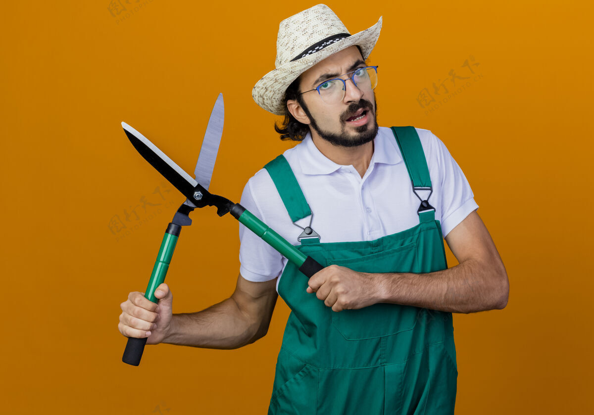 人年轻的留着胡子的园丁 穿着连体衣 戴着帽子 拿着树篱剪 站在橘色的墙上不高兴地看着前面姿势站着衣服