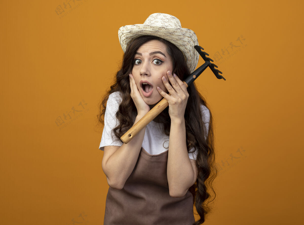 人震惊的年轻女园丁穿着制服戴着园艺帽把手放在脸上拿着耙子隔离在橙色的墙上人手人