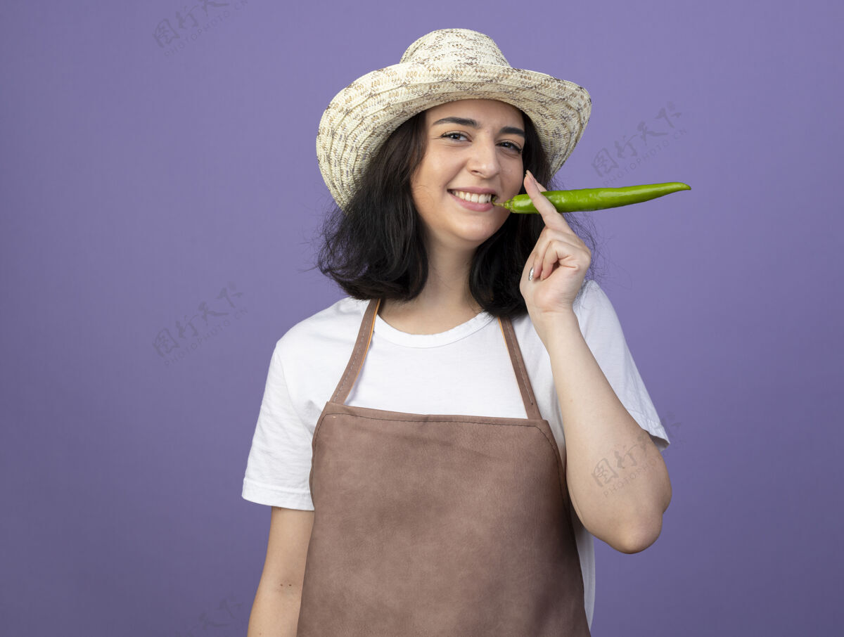 嘴微笑着的年轻黑发女园丁穿着制服 戴着园艺帽 紧握着辣椒的嘴 隔离在紫色的墙上胡椒拿着人