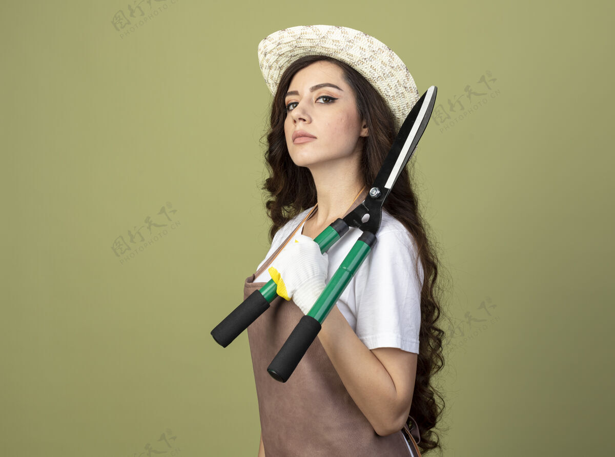 穿自信的年轻女园丁穿着制服 戴着园艺帽和手套 站在一旁 肩上扛着园艺剪 隔离在橄榄绿的墙上女公民人