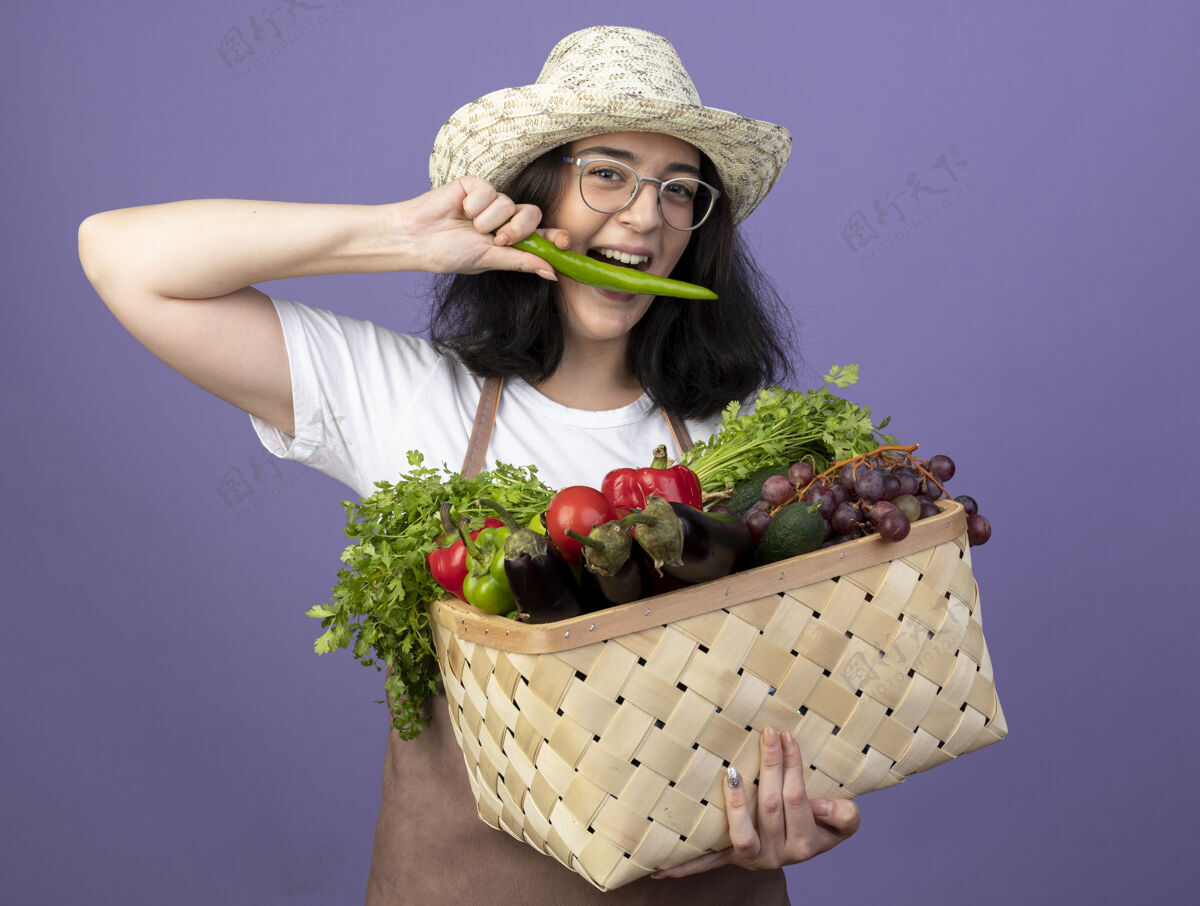 市民戴着眼镜 穿着制服 头戴园艺帽的快乐的年轻黑发女园丁 手里拿着菜篮子 假装咬着隔离在紫色墙上的辣椒年轻的制服假装