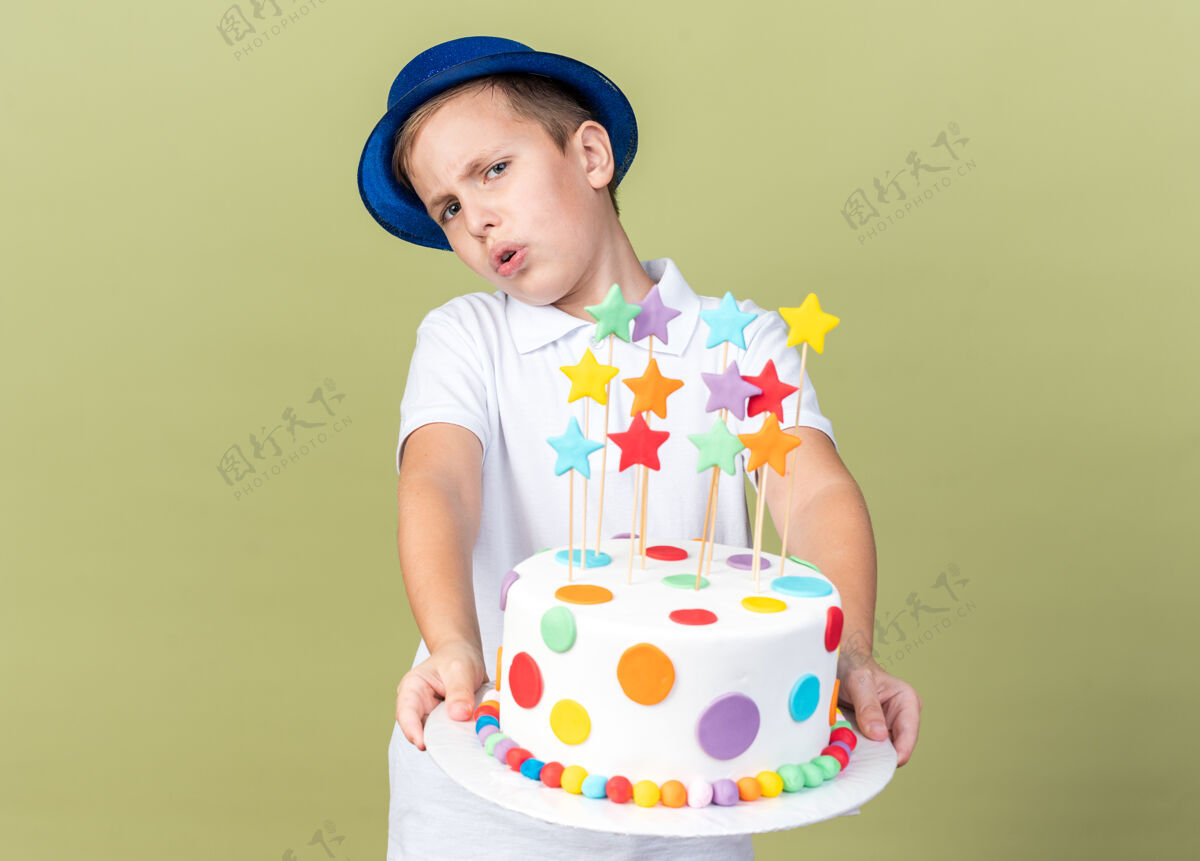 斯拉夫恼怒的年轻斯拉夫男孩 戴着蓝色派对帽 拿着生日蛋糕 隔离在橄榄绿的墙上 留着复印空间帽子孤立年轻