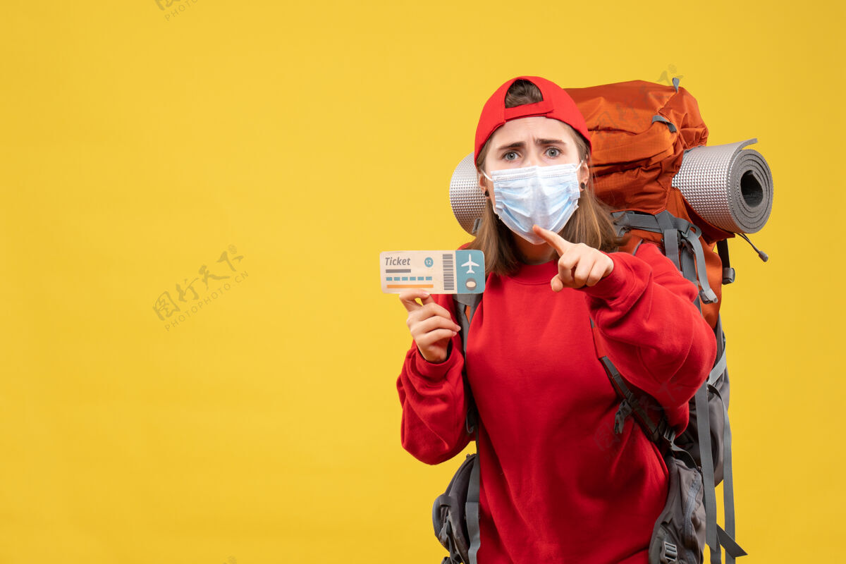 票正面图年轻的女旅客背着背包 戴着口罩拿着票 手指朝上拿着指着面具