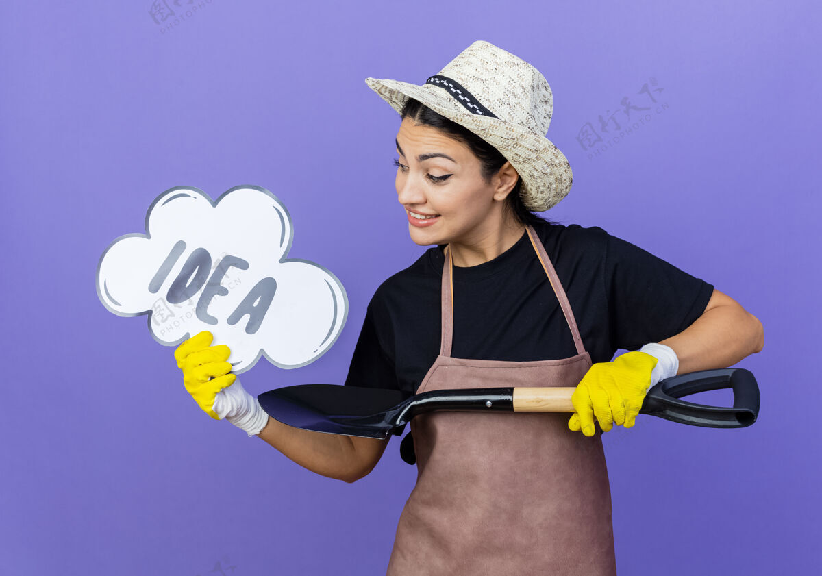 微笑年轻漂亮的女园丁穿着围裙 戴着帽子 拿着铲子 站在蓝色的墙上说着“创意”这个词花园人人