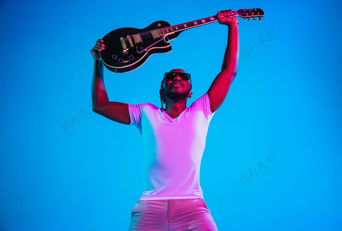 艺术家年轻的非洲裔美国音乐家在霓虹灯下像摇滚明星一样在蓝色的墙上弹吉他男人球员成人