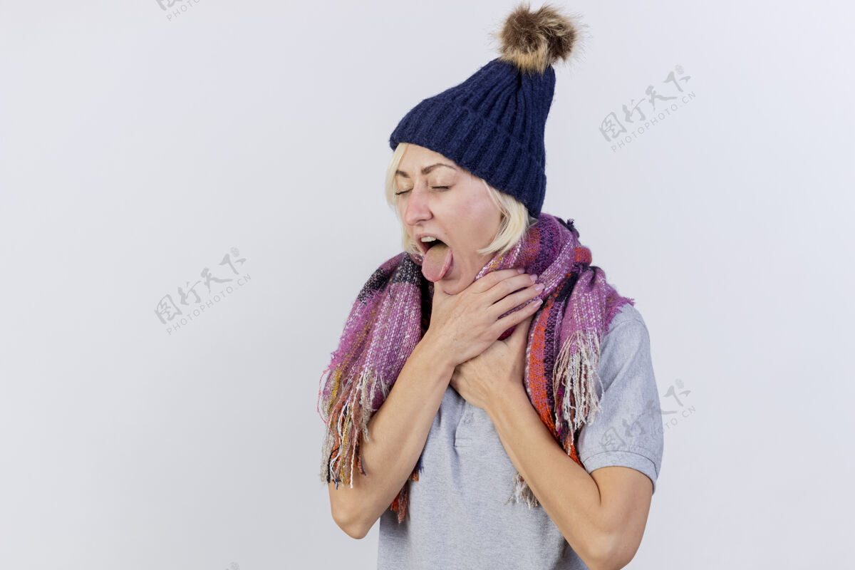 姿势恼怒的年轻金发病女人戴着冬天的帽子和围巾结结巴巴的舌头 把手放在脖子上隔离在白色的墙上斯拉夫年轻人疾病