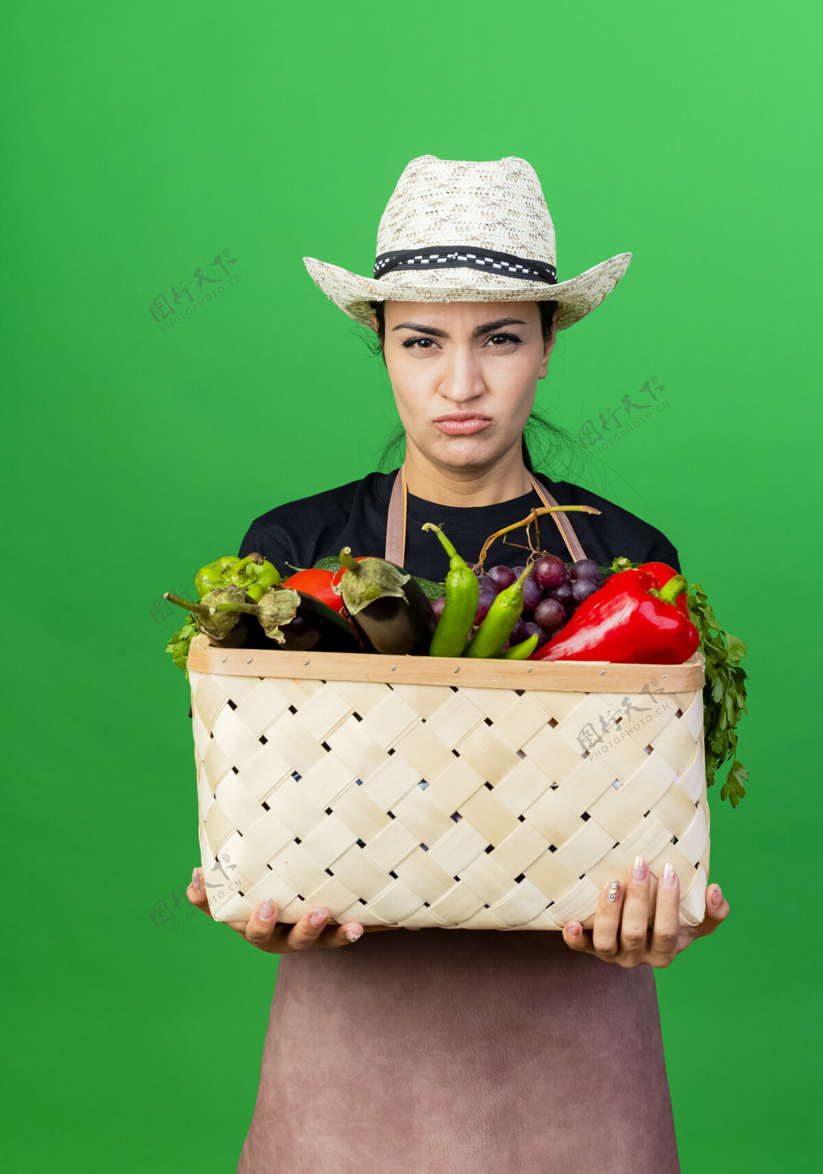 市民年轻漂亮的女园丁 围着围裙 戴着帽子 手里拿着装满蔬菜的篮子 站在绿色的墙上 严肃地看着前面 脸上露出不高兴的神情人人花园