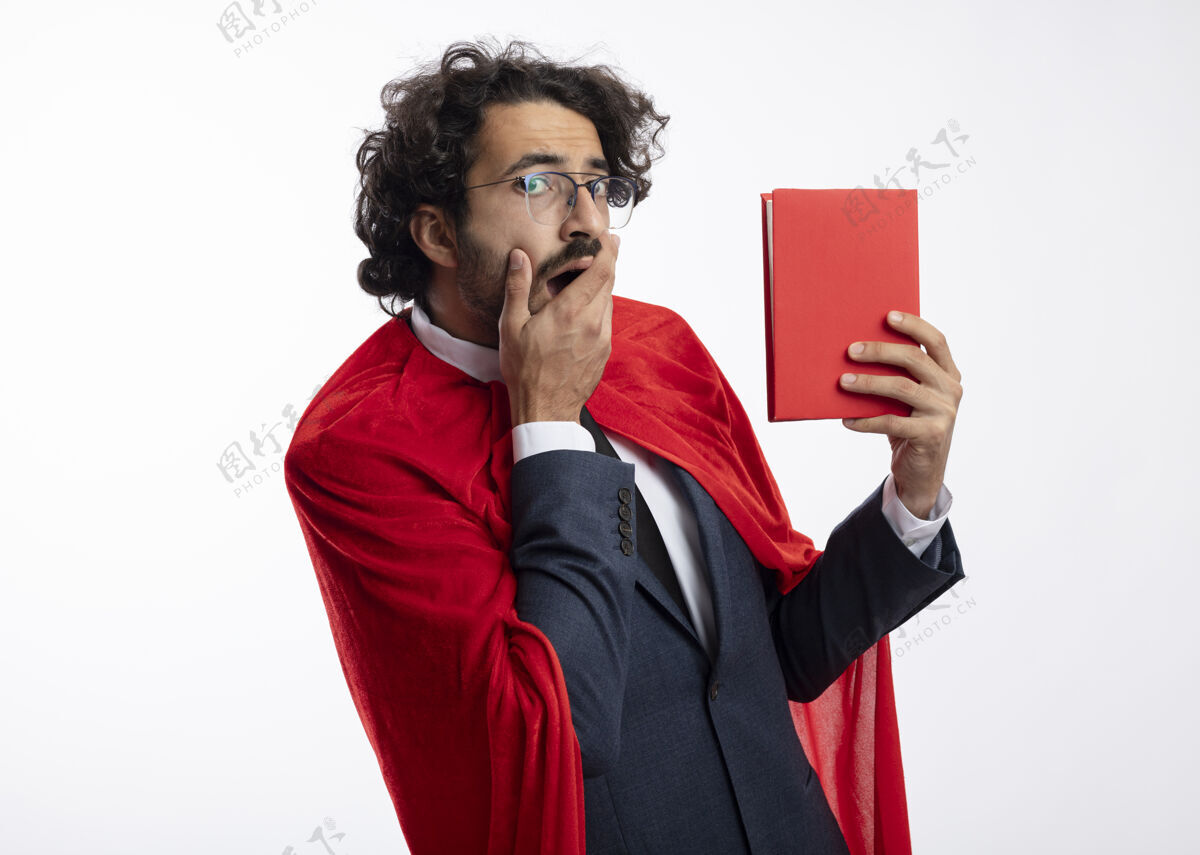 表情兴奋的年轻超级英雄戴着眼镜 穿着红色斗篷的西装 手放在嘴上 把书孤立地放在白墙上年轻西装脸