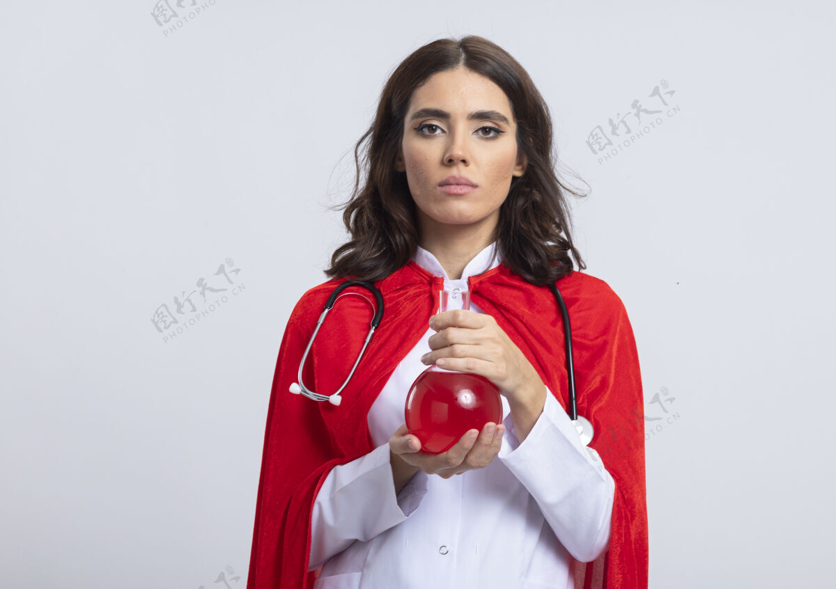 脸自信的女超人穿着医生制服 戴着红色斗篷 听诊器拿着隔离在白墙上的玻璃瓶中的红色化学液体制服姿势女孩
