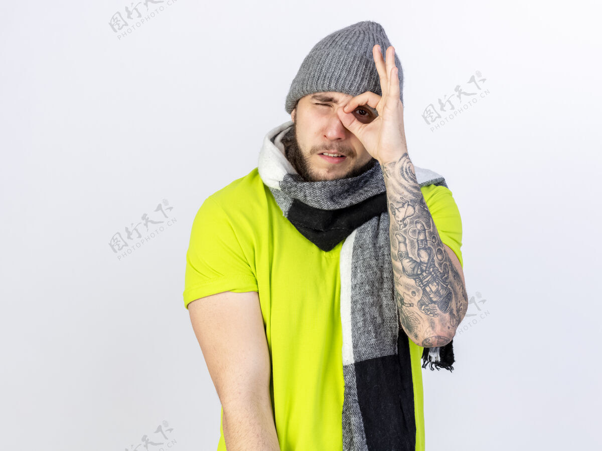 年轻人戴着冬天帽子和围巾的自信的年轻病夫隔着白墙上的手指望着前方人穿人