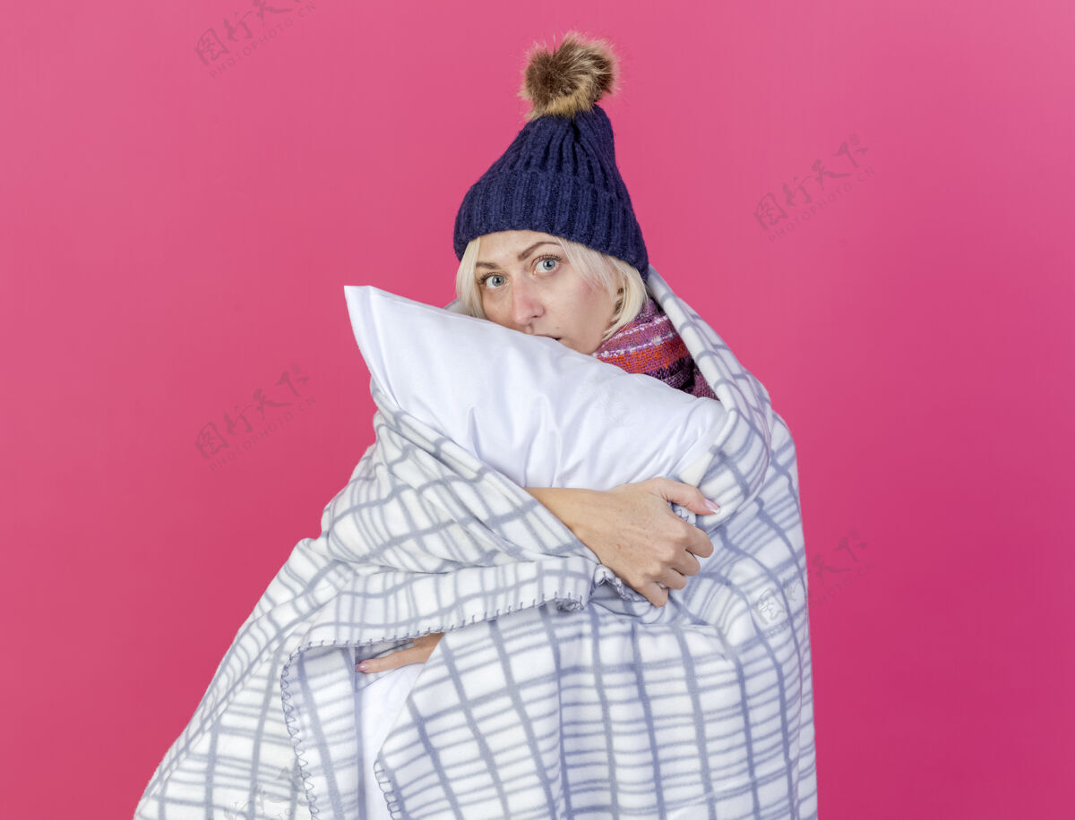 姿势焦急的年轻金发病女人戴着冬天的帽子和围巾裹着格子呢抱枕看着前面孤立的粉色墙壁围巾帽子人