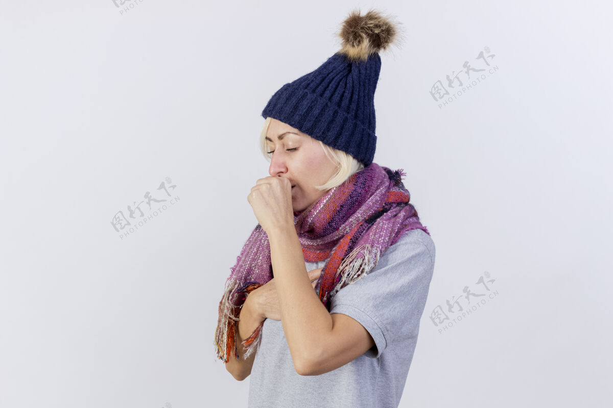 脸咳嗽的年轻金发病女人戴着冬天的帽子和围巾 紧握着拳头 嘴巴被隔离在白色的墙上帽子人穿