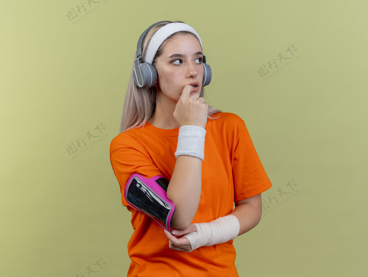 困惑困惑的年轻运动女性戴着耳机戴着牙套戴着头带腕带和电话臂章手放在下巴上看着橄榄绿墙上孤立的一面电话头带运动