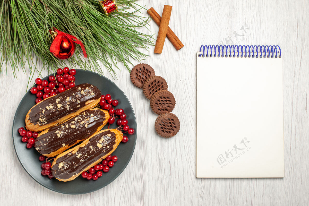 肉桂俯瞰图：灰色盘子上的巧克力和葡萄干饼干交叉着肉桂和松树叶子 白色木桌上放着圣诞玩具和笔记本圣诞烹饪玩具
