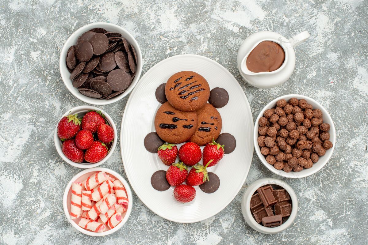 餐白色椭圆形盘子上的巧克力饼干草莓和圆形巧克力 灰白色地面上的碗里放着糖果草莓巧克力麦片和可可食物浆果早餐