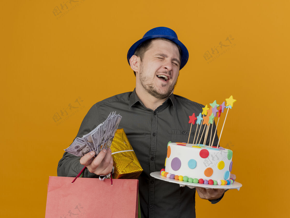 派对快乐与闭上眼睛的年轻人戴着蓝色的帽子拿着蛋糕与礼物和现金孤立在橙色聚会家伙穿礼物