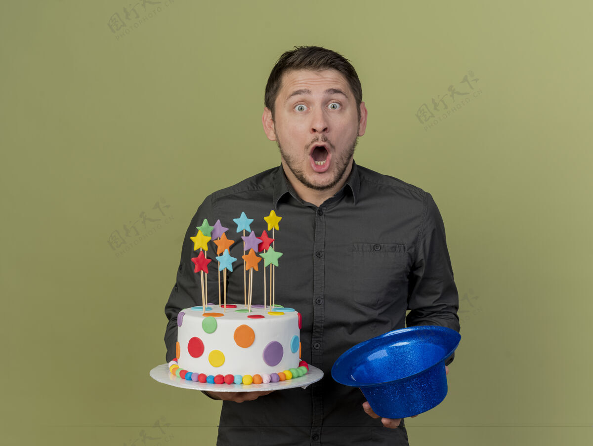 蓝色惊讶的年轻人穿着黑色衬衫拿着蛋糕和蓝色帽子隔离在橄榄绿惊喜派对小伙子