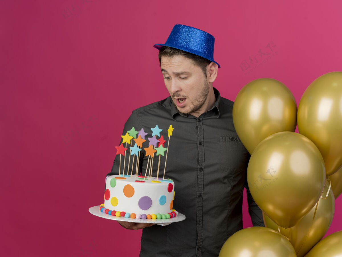 年轻人一个戴着蓝色帽子的年轻人站在气球旁边 拿着一个粉红色的蛋糕看着蓝色穿上帽子