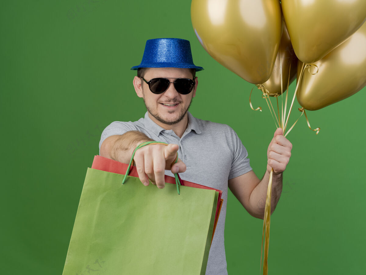 举行微笑的年轻人戴着聚会帽和眼镜拿着气球和礼品袋 向你展示绿色的手势眼镜礼物绿色