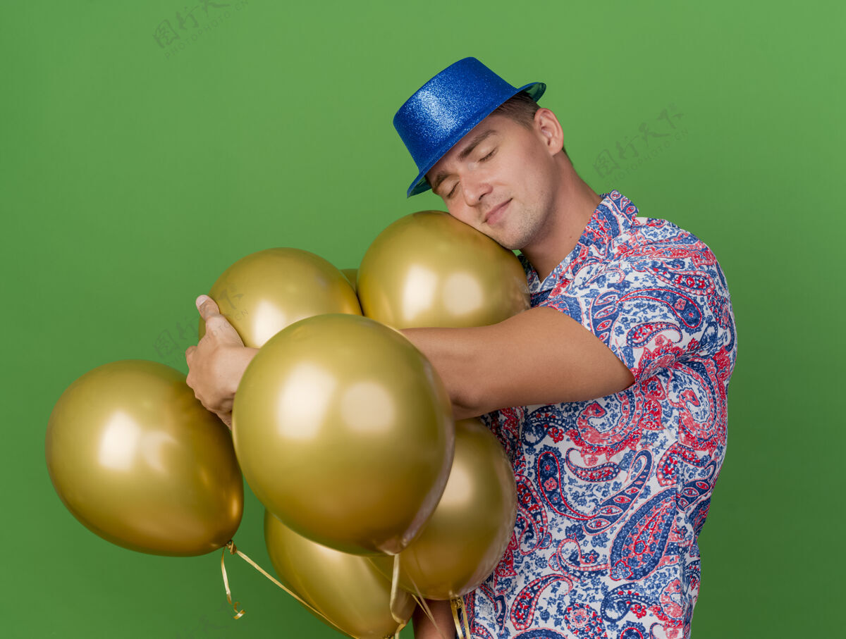 派对戴着蓝帽子闭着眼睛的年轻人站在绿色的气球旁年轻蓝色伙计
