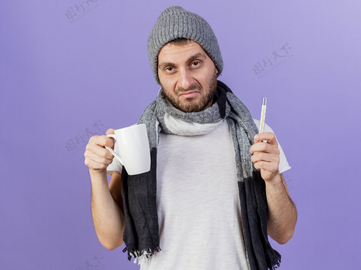 悲伤悲伤的年轻人戴着冬天的帽子 戴着围巾 拿着温度计 把一杯紫色的茶隔上男人茶围巾