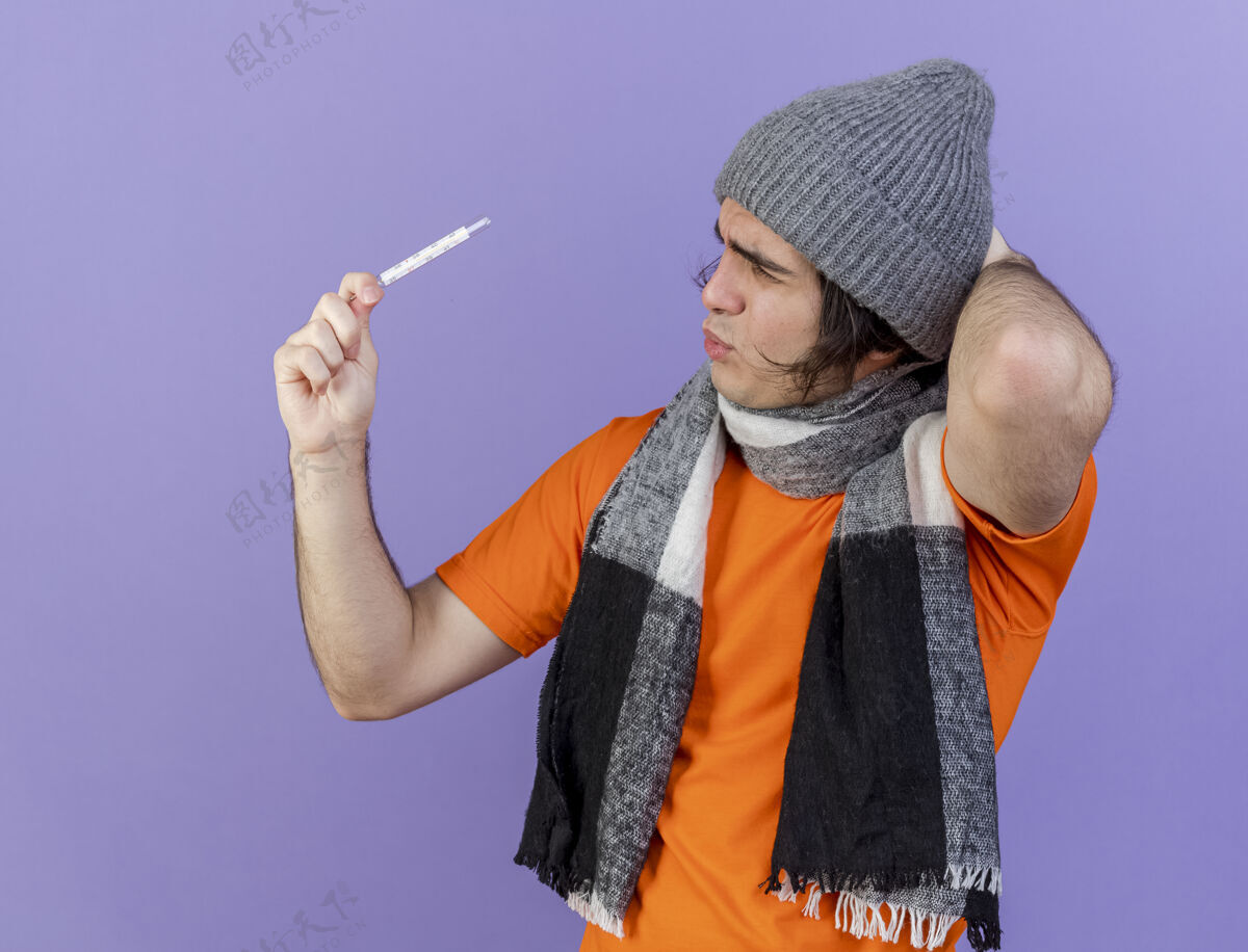 疾病困惑的年轻病人戴着冬天的帽子 戴着围巾 看着温度计 把手放在脑后 孤立在紫色的头上男人帽子穿