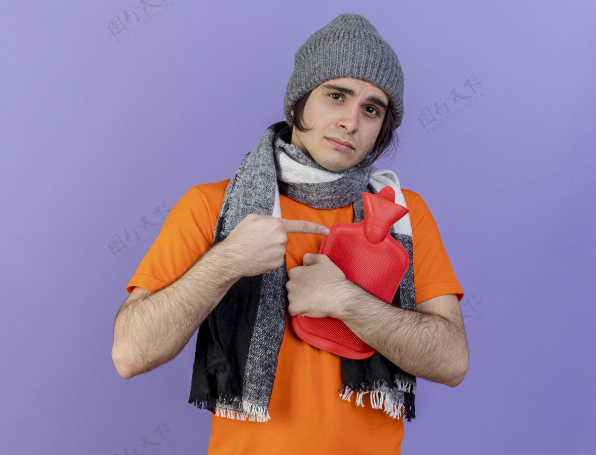 穿一个戴着冬天帽子 戴着围巾的年轻人拥抱着 指着紫色的热水袋拥抱帽子包