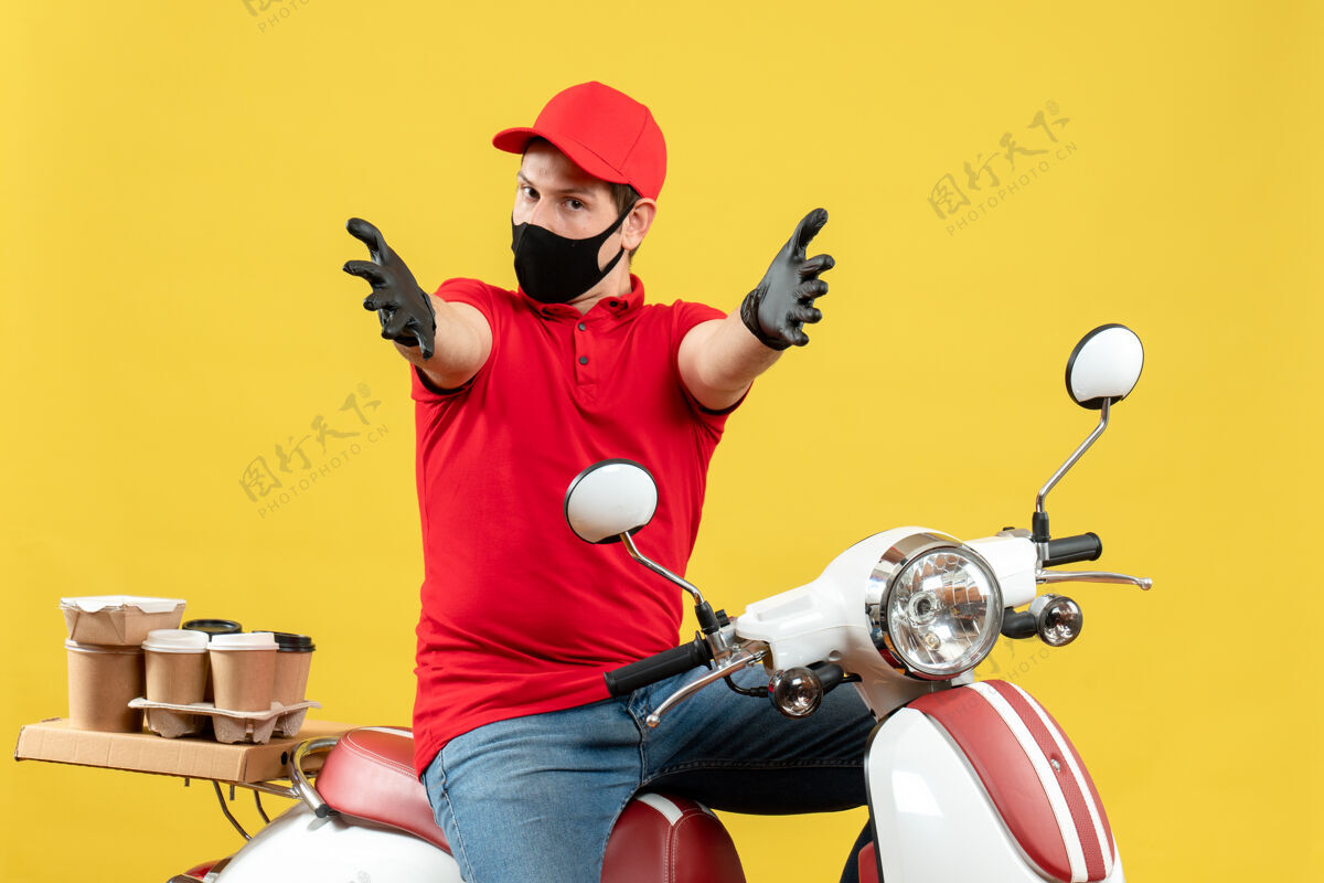 手臂俯视图：戴着医用口罩 穿着红色上衣 戴着帽子手套的年轻人坐在滑板车上 感觉困惑 在黄色背景下向前伸展双臂困惑秩序帽子
