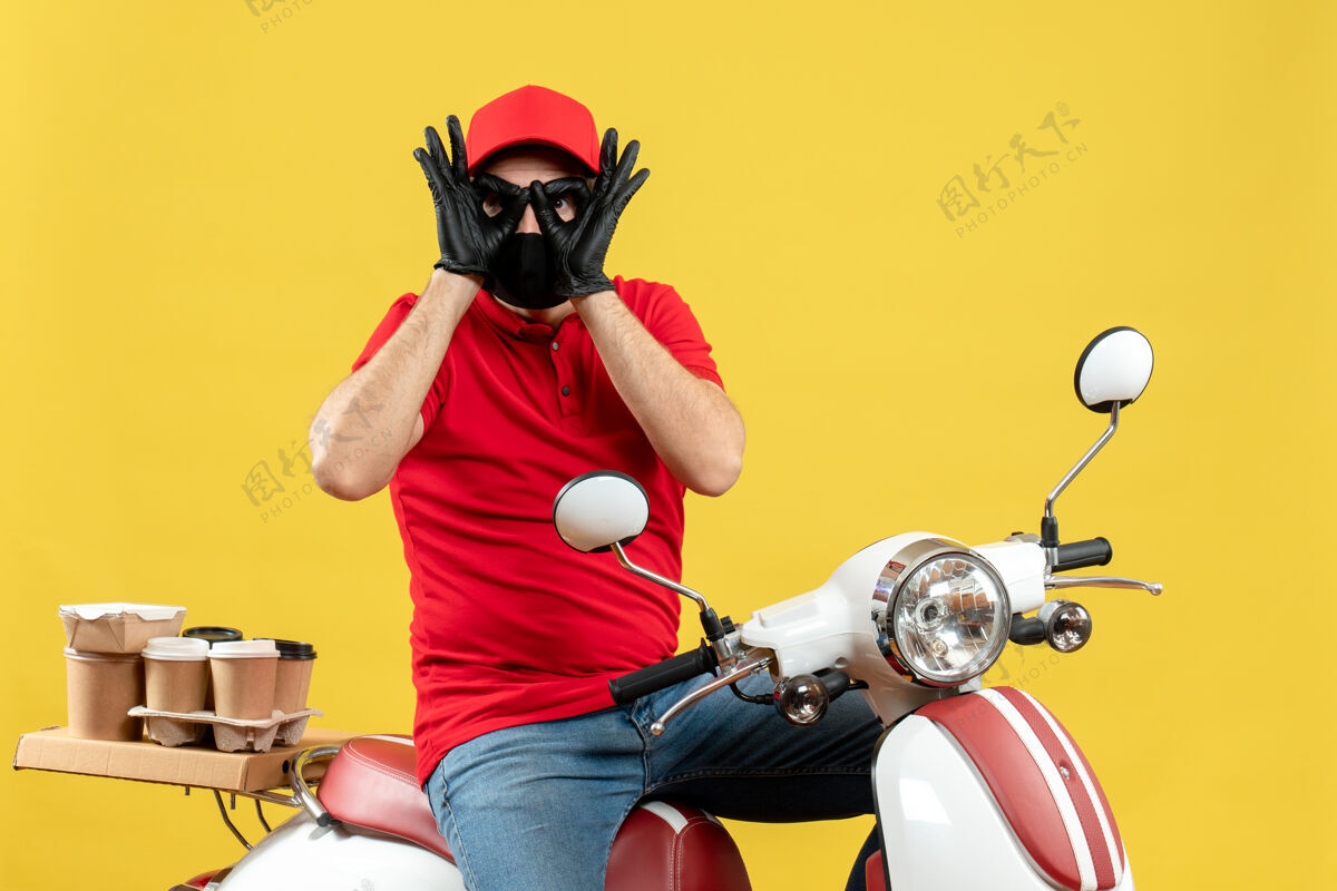 摩托车戴着医用口罩 穿着红色上衣 戴着帽子手套的年轻人的俯视图传达命令坐在踏板车上 在黄色背景上做着戴眼镜的手势面罩医疗背景